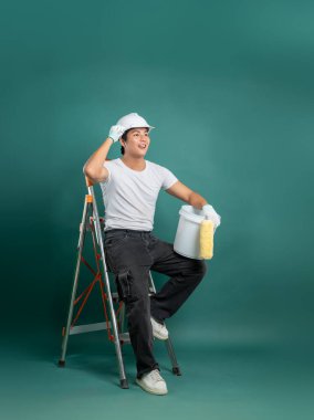 Beyaz tişörtlü ve koyu renk kotlu genç bir ressam, sert bir şapka takmış, elinde boya silindiri tutarak yeşil bir zemine karşı bir merdivene oturur. Yukarı ve uzağa bakıyor..