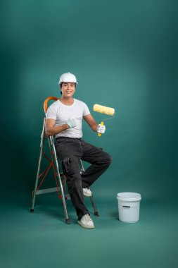 Bir ressam merdivene elinde bir silindirle oturur ve gülümser. Sert bir şapka takıyor ve yeşil bir arka fona karşı bir kovanın yanındaki patenciye işaret ediyor. Ev geliştirme.