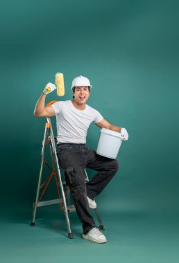 Bir ressam, elinde boya rulosu ve kova tutarak sevinçle bir merdivene poz verir, deniz mavisi arkaplanına dayanır, ev yapımı veya DIY projeleri için idealdir.. 