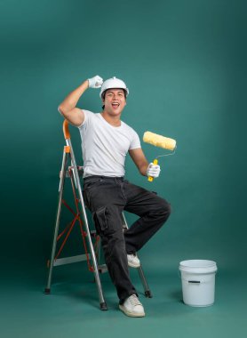 Beyaz miğferli ve eldivenli bir ressam, elinde boya silindiri, esneme ve boya malzemeleriyle yeşil bir zemine neşe saçan bir merdiven üzerinde oturur..