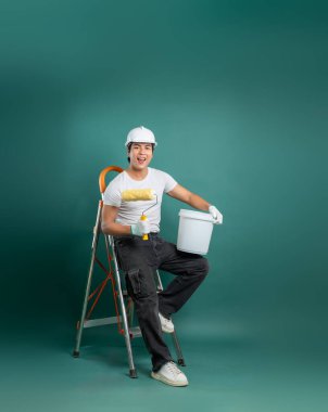 Koruyucu kıyafet giyen tutkulu bir ressam elinde silindir olan bir merdivende oturur ve sağlam yeşil bir zemine karşı boya kovası çizer. Ev geliştirmeye hazır..