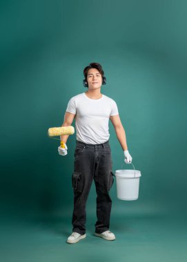 Kulaklık takan bir ressam, bir elinde boya silindiri, diğer elinde boya kovası, yeşil arka planda..