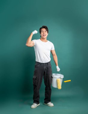 Beyaz tişörtlü, siyah pantolonlu, beyaz ayakkabılı ve eldivenli genç bir adam bir kova tutarken kolunu esnetiyor ve koyu yeşil arka planda boya rulosu yapıyor..