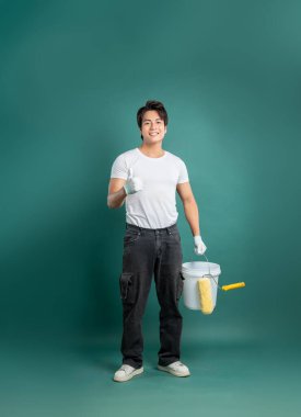 Ressam, yeşil arka planda duran koruyucu eldivenlerle ve kovayla çalışmaya hazır. Başparmağını kaldırıyor ve gülümsüyor.