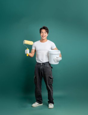 Beyaz tişörtlü ve kargo pantolonlu gülümseyen bir ressam boyamaya hazır. Elinde bir boya silindiri ve bir kova tutuyor yeşil bir arka fona karşı.