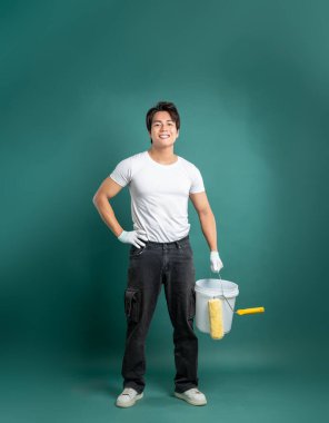 Kovası ve silindiri olan dost canlısı genç bir ressam sağlam bir arka plana karşı duruyor. Ev yapımı bir projeyle uğraşmaya hazır, yeni bir kat boya getiriyor..