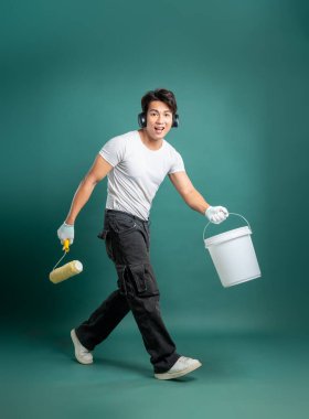 Kulaklıklı gülümseyen bir adam elinde boya silindiri ve kova tutuyor. Ev yenileme projesine başlamaya hazır. Üzerinde eldiven ve iş kıyafetleri var..