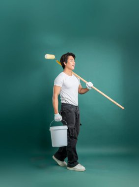 Omzunda boya rulosu olan, elinde boya kovası olan, eldiven takan, yeşil arka planlı bir stüdyoda duran, sağ tarafa bakan bir ressam..