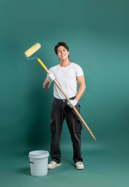 Gülümseyen bir adam elinde boya silindiri ve kovayla güvenli bir şekilde duruyor ve DIY ev geliştirme projesine başlamaya hazır. Beyaz bir gömlek ve yeşil bir arka plana karşı günlük kıyafetler giyer..