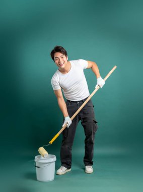Beyaz tişörtlü ve siyah pantolonlu gülümseyen bir adam deniz mavisi arkaplanının önünde boyamaya hazır bekliyor. Elinde boya silindiri ve bir kova var, DIY projesi için poz veriyor..