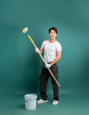 Eldivenli ve işçi pantolonlu gülümseyen bir tamirci yeşil arka planda boya silindiri ve kova tutar, boyama işleri için hazırdır, DIY veya yenileme projeleri için uygundur.