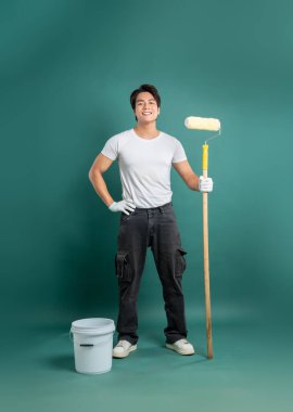 Gülümseyen bir ressam güvenle bir deniz mavisi arkaplanına karşı durmaktadır. Beyaz tişört, siyah iş pantolonu ve eldiven giyiyor. Elinde bir kova dolusu boya silindiri tutuyor..