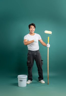 Yetişkin bir adam duvarları boyamaya hazır, yanında kova olan bir boya silindiri ile yeşil bir zemin üzerinde duruyor, bu da bir DIY yenileme projesini gösteriyor..