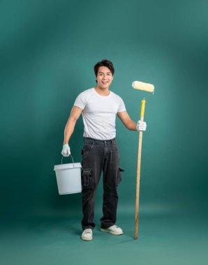 Gülümseyen bir insan elinde boya rulosu ve kovayla resim yapmaya hazırdır. İş kıyafetleri içinde, ev gelişimini, DIY görevlerini veya genel kasılmaları yakalar..