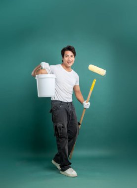 Beyaz gömlekli ve eldivenli gülümseyen bir ressam, elinde bir boya kovası ve yeşil bir arka fona karşı bir silindir tutuyor. Ev boyama işi için hazır..