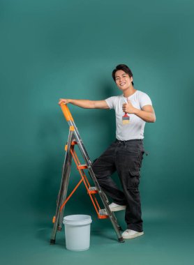 Elinde boya kovası ve fırçayla merdivende gülümseyen bir ressam, yeşil bir zemine yaslanmış, resme hazır bir şekilde baş parmak hareketi yapıyor..
