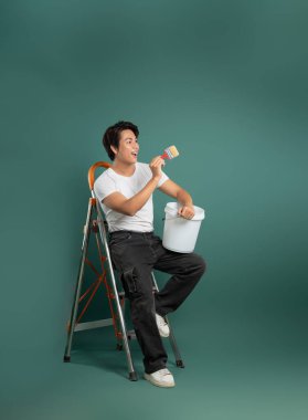 Sıradan kıyafetlerle merdivende oturan, elinde boya fırçası ve kovayla yeşil bir zemine gülümseyen genç adam..