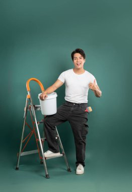 Beyaz gömlekli ve siyah pantolonlu gülümseyen bir adam elinde boya kovası olan bir merdivende duruyor, yeşil bir arka plana başparmak işareti veriyor, boyamaya hazır..