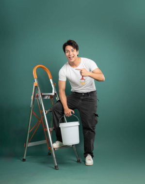 Elinde boya kovası olan neşeli bir adam gömleğindeki boya fırçası tasarımını esprili bir şekilde işaret ederek bir resim projesine eğlenceli bir yaklaşım öneriyor..