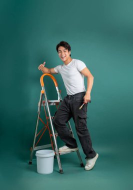 İş kıyafetleri içinde neşeli bir adam boya fırçasıyla merdivene dayanıyor ve başparmağını kaldırıyor. Deniz mavisi arkaplanına ve yakınlardaki kovaya resim yapmaya hazır..