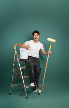 Neşeli bir adam boya silindiri ve kovayla merdivenin üzerinde duruyor, gülümsüyor ve başparmağını kaldırıyor. Stüdyo çekimi yeşil zemin üzerinde. DIY, ev onarımı ve boyama.