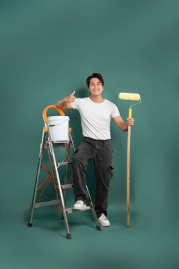 Gülümseyen bir insan kendine güvenen bir boya silindiri ile ayakta duruyor, bir stüdyo arka planında elinde boya kovası olan bir merdivenin yanında başparmak işareti veriyor. Ev projesi.