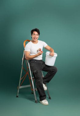 Gülümseyen genç bir adam elinde boya fırçası ve kovayla merdivende oturuyor, odayı boyamaya hazır. Günlük kıyafetler ve beyaz spor ayakkabılar giyiyor.. 