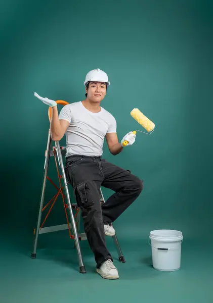 Beyaz gömlekli, kasketli ve eldivenli rahat bir işçi boya silindirli bir merdivende oturuyor. Beyaz bir boya kovası yeşil arka planda yerde duruyor..
