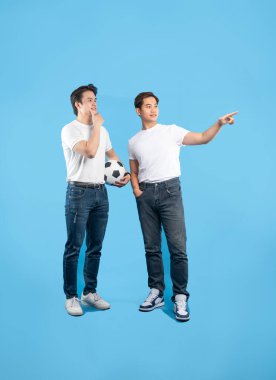 Two young men in jeans and t-shirts stand against a light blue background. One holds a soccer ball while pondering, the other points towards the distance. clipart