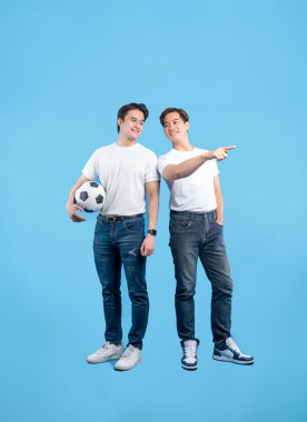 İki sıradan giyimli genç adam açık mavi bir arkaplana karşı poz veriyor. Biri bir futbol topu tutarken, diğeri yan tarafı işaret ediyor. İkisi de gülümsüyor..
