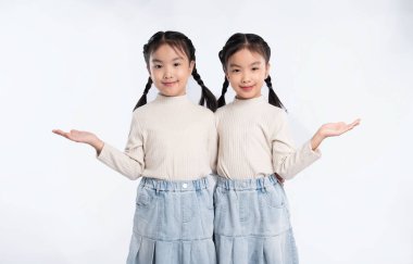 İki genç ikiz kız omuz omuza, beyaz bir arka planda, at kuyruğu ve benzer kıyafetlerle, avuç içleri boşluğa bakıyor..