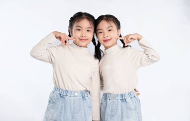 İki genç, tek yumurta ikizi kız fotoğraf için poz veriyor. Birbirlerine uyumlu kıyafetler giyiyorlar, gülümsüyorlar, elleri yanaklarında, beyazlara karşı duruyorlar..