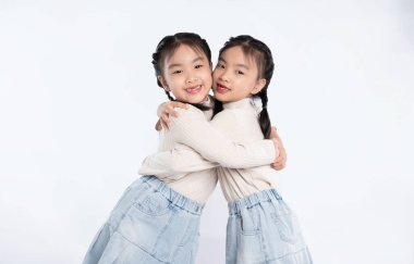 Tek yumurta ikizleri mutlu bir şekilde birbirlerine sarılıyorlar, üst ve etek giyiyorlar. Temiz, beyaz bir arkaplanda konumlanmışlar, bağlantılarını vurguluyorlar..