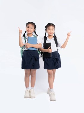 Aynı üniformayı giyen iki genç kız ellerinde kitaplarla beyaz bir arkaplanda duruyorlar. Sırt çantaları taşıyorlar. Heyecan ve beklenti sinyalleri veriyorlar..