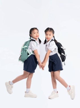 Okul üniformalı iki genç kız el ele tutuşup sırt çantalarıyla beyaz bir zemine yaslanıyorlar. Okul için hazır görünüyorlar..