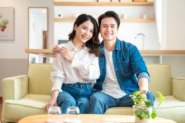 Açık yeşil bir koltukta oturan mutlu bir Asyalı çift parlak, modern bir oturma odasında gülümsüyor.