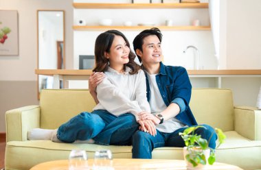 Mutlu genç bir çift, modern güneşli dairelerinde sarı bir kanepede birlikte oturur ve nazikçe gülümserler.