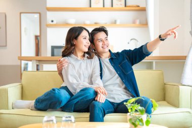 Gülümseyen genç bir çift, gündelik kıyafetlerini giymiş, parlak ve modern bir yaşam alanında koltukta dinleniyorlar.