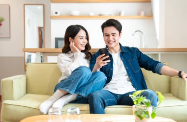 Çift, geniş bir pencereden gelen ışıkla birlikte modern bir oturma odasında dinlenerek ekranda vakit geçirmeyi seviyor.