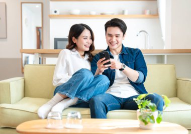 Gülümseyen bir çift, modern oturma odasındaki açık yeşil bir kanepede dinlenirken aynı telefonu paylaşıyor.