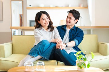 Genç bir Asyalı çift sarı bir kanepede oturmuş el ele tutuşuyor ve gülümsüyor.