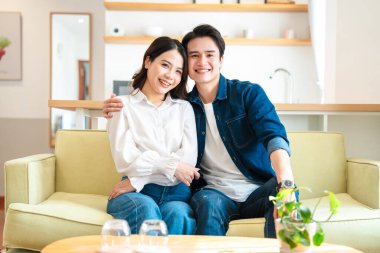 Gülümseyen Asyalı bir çift gündelik kıyafetleriyle modern apartmanlarında bir kanepeye sarılıyor.