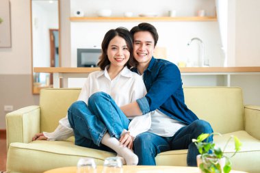 Asyalı bir çift, modern ve minimalist bir evde koltuklarında dinlenirken gülümsüyor.