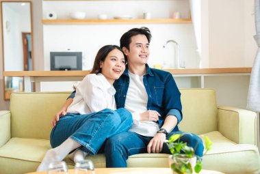 Asyalı çift koltukta rahat rahat gündelik giysiler içinde modern apartman dairesinde birlikte vakit geçiriyorlar.