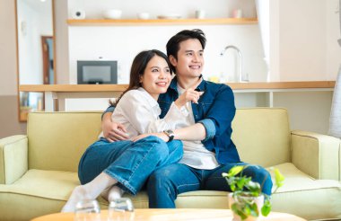 Gülümseyen bir çift kanepede oturup uzaktan bir şeye bakar, işaret eder ve kucaklaşır.