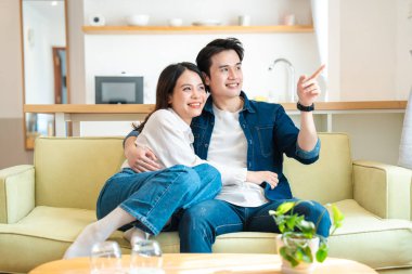 Gülümseyen Asyalı çift modern bir dairede koltukta oturuyor.