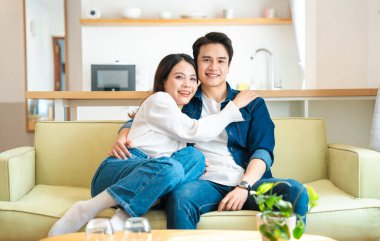 Bir çift kanepeye yakın oturuyor. Gülümsüyor ve kameraya bakıyor. Rahatlamış görünüyorlar.