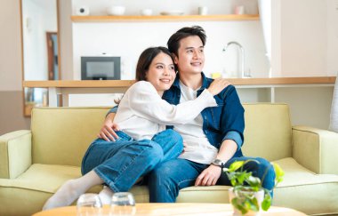 Genç bir çift, modern ışıkla dolu bir evde sarı bir kanepeye sarılır ve gülümseyen bir şekilde pencereden dışarı bakar.