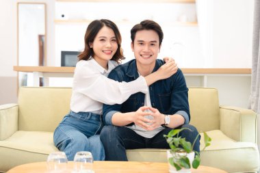 Gülümseyen Asyalı bir çift, modern, parlak görünümlü bir evde, bir kanepede birlikte oturuyor.