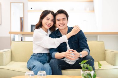 Mutlu bir çift, parlak ve serin oturma odalarında kanepede oturup kucaklaşır ve gülümserler.